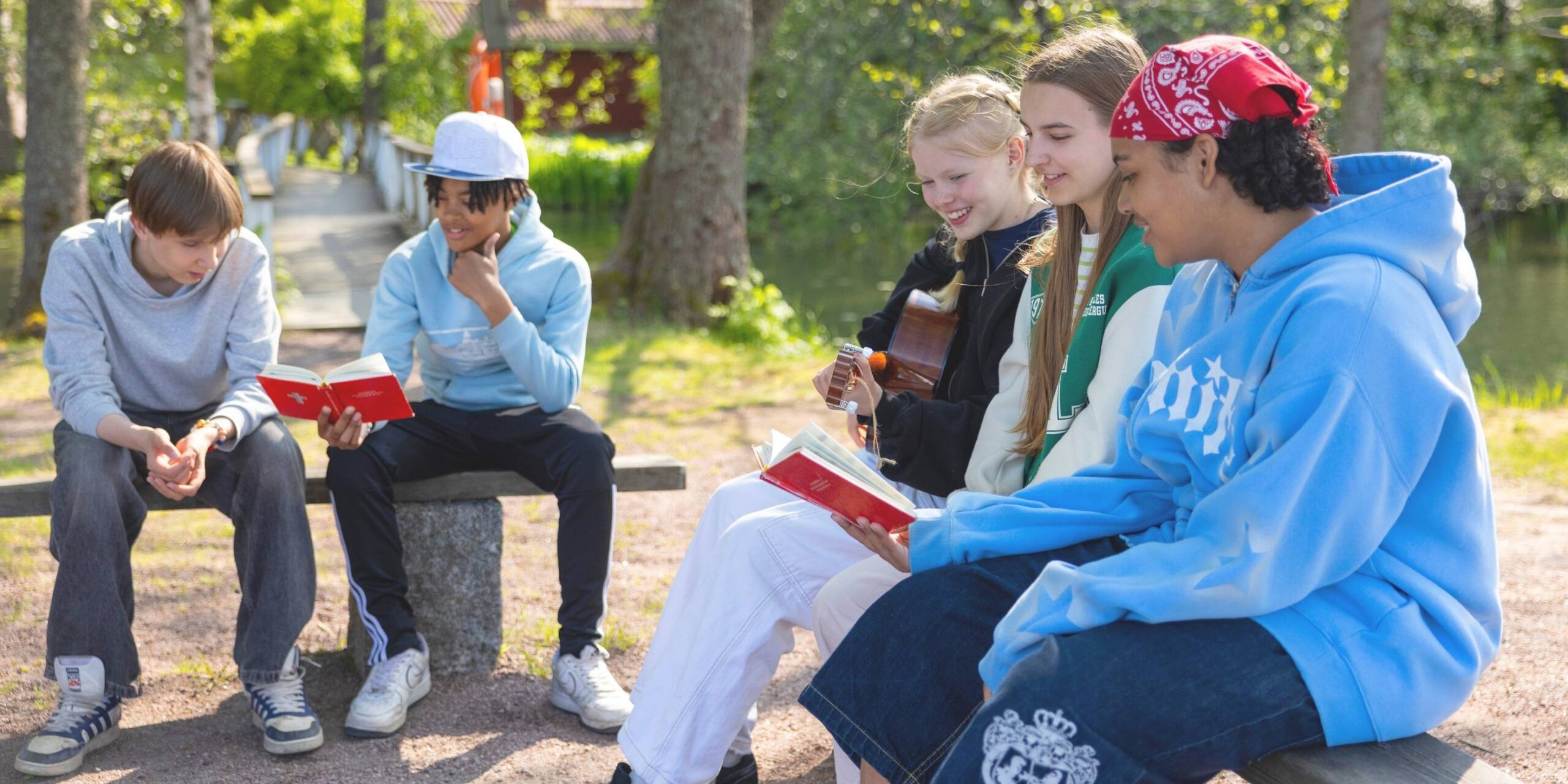 Nuoria ystäviä istumassa penkillä, lukemassa kirjoja ja soittamassa kitaraa kauniissa kesäisessä ympäristössä. Taustalla puut ja järvi.