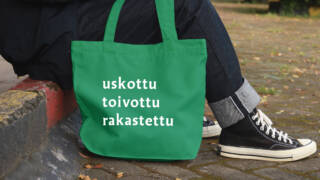 Maahan on laskettu kangaskassi, jossa lukee uskottu toivottu rakastettu. Ihminen istuu portailla kassin kanssa.
