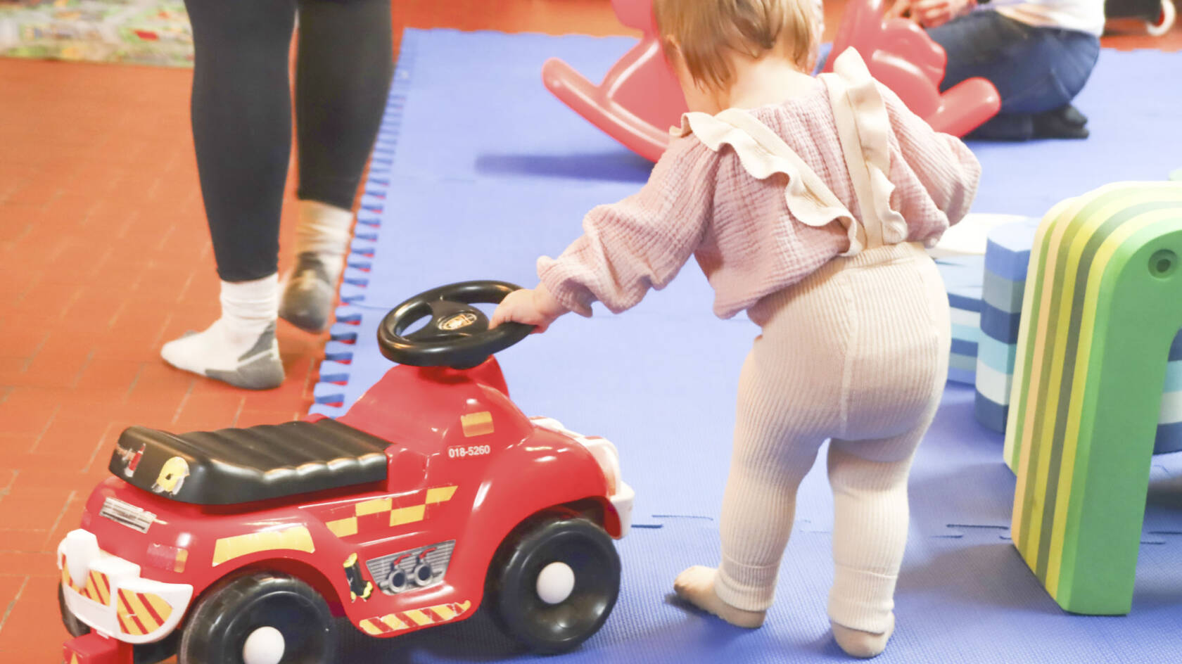 Pieni taapero selin kuvaan päin. Taaperolla punainen potkuauto.