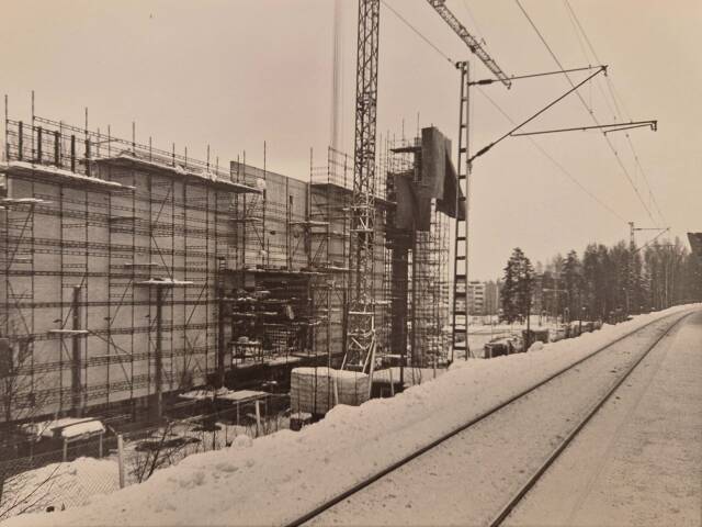 Mustavalkoinen kuva, etualalla junaraiteet, taustalla iso rakennus rakennustelineiden keskellä ja nosturi.