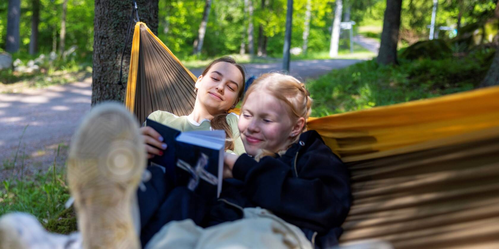 Kaksi nuorta naista makaa riippumatossa metsässä, lukien kirjaa ja nauraen yhdessä. Taustalla vihreitä puita ja polku.