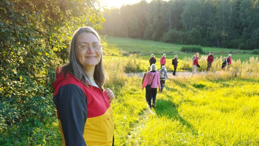 nainen aurinkoisen niityn reunalla, taustalla retkeilijöitä jonossa